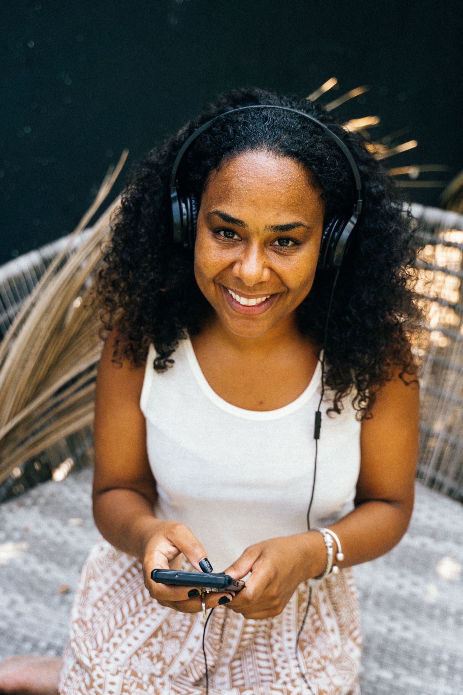 Smiling young Black woman with curly hair wearing headphones, holding her phone in both hands and looking up at the camera
